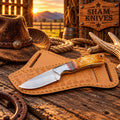 D2 cowboy skinner knife with a burnt bone handle resting on a tan tooled leather sheath

