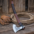 Handmade Viking axe with engraved carbon steel blade and leather wrapped wooden handle on rustic barn table with rope and gloves